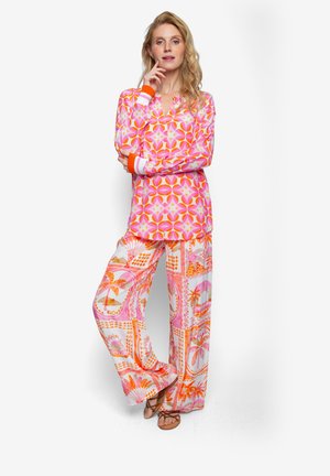 Różowa bluzka z długim rękawem z geometrycznymi wzorami i pomarańczowymi mankietami w paski, zestawiona z szerokimi spodniami o żywych tropikalnych wzorach i kolorach.