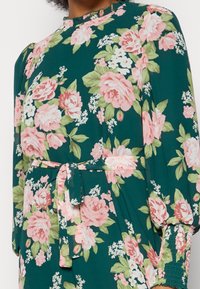 Vestido floral en verde oscuro con rosas rosas y acentos blancos. Presenta un cuello alto, mangas gather y un cinturón atado en la cintura. Tela suave.