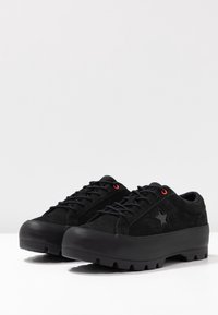 Para czarnych sneakersów na platformie z grubymi podeszwami, detalem gwiazdy po bokach i czerwonymi oczkami przy sznurowadłach, na białej powierzchni.