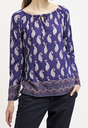 Vrouw draagt een blauw blouse met lange mouwen en witte paisleyprints en een patroonzoom, gecombineerd met zwarte broek, hand in de zak.