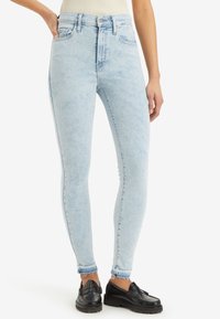 Jeans skinny a vita alta blu chiaro con un lavaggio sbiadito, caratterizzati da un classico design a cinque tasche e orlo sfilacciato, abbinati a mocassini neri.