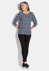 Blaue und lila gemusterte Dreiviertelarmbluse, runder Ausschnitt, kombiniert mit schwarzen Skinny Jeans und schwarzen Sneakers. Glatte Stofftextur.