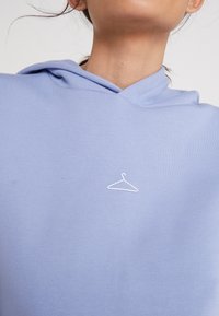 Sudadera azul claro fabricada con un material suave, con capucha ajustable. Presenta un logo de percha bordado en blanco en el pecho.