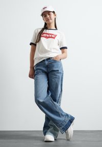 White t-shirt with red Levi's logo and navy trim, paired with wide blue denim jeans and white sneakers. Model wears a white cap.