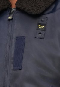 Veste bleu marine avec un col en shearling noir doux, dotée d'un patch logo, d'une fermeture éclair et d'un tissu texturé avec un détail de sangle verticale.