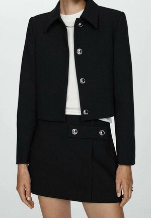 Zwarte cropped jas met zilveren knopen over wit topje, gecombineerd met een bijpassende zwarte minirok met knoopdetails aan de tailleband.