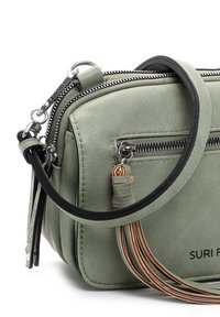 Zielona torba z ekologicznej skóry z dwoma zamykanymi na zamek komorami, czarną rączką oraz ozdobnym chwostem z brązowych i beżowych sznurków. Posiada logo.