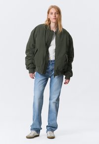 Jeune personne debout, portant une grande veste bomber vert foncé, un t-shirt blanc, un jean bleu clair et des baskets blanches, sur un fond blanc uni.