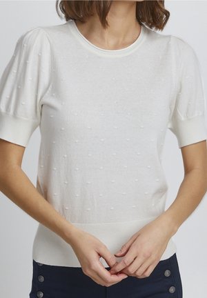 Frau trägt einen weißen, strukturierten Kurzarm-Pullover mit Puffärmeln und marineblaue Hose mit Knopfdetails.