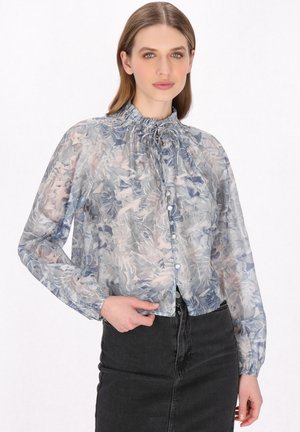 Blusa trasparente a maniche lunghe in tonalità blu chiaro e rosa con motivo floreale. Presenta un collo arricciato e una chiusura con bottoni, indossata sopra una gonna di jeans scura.