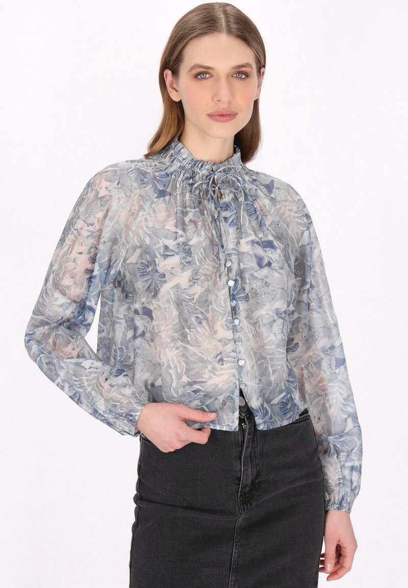Hame baju lengan panjang yang transparan dengan pola bunga biru muda dan merah jambu. Memiliki leher berenda dan kancing di bagian depan, dikenakan di atas rok denim gelap.