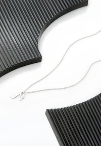 Sølv halskæde med en musiknote vedhæng, sat mod strukturerede sorte buede objekter. Kæden er delikat med et enkelt design.