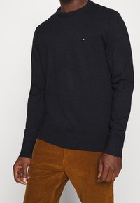 Un homme portant un pull noir à manches longues avec un petit logo sur la poitrine et un pantalon en velours côtelé marron, se tenant devant un arrière-plan uni.