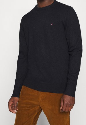 Mann in einem schwarzen Langarmpullover mit einem kleinen Logo auf der Brust und braunen Cordhosen, der vor einem schlichten Hintergrund steht.