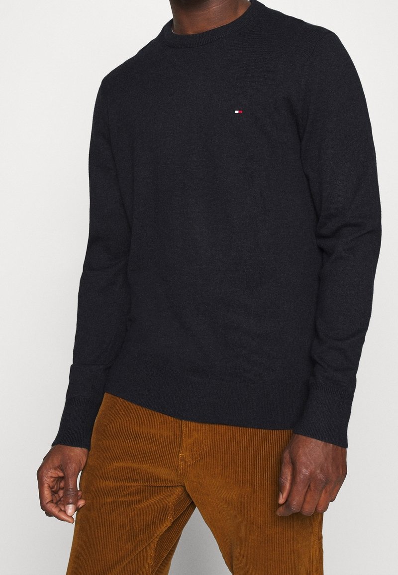 Un homme portant un pull noir à manches longues avec un petit logo sur la poitrine et un pantalon en velours côtelé marron, se tenant devant un arrière-plan uni.