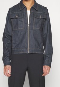 Chaqueta de mezclilla azul oscuro con cremallera frontal, bolsillos en el pecho con botón, llevada sobre una blusa blanca y combinada con pantalones negros.