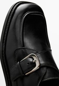 Zwarte leren schoen met een afgeronde neus, strak ontwerp, decoratieve gespsluiting en contrasterende stiksels langs de naad.