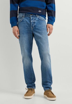 Man met lichtblauwe spijkerbroek, bruine veterschoenen en een blauw-zwart gestreept shirt met lange mouwen, staand op een lichte achtergrond.
