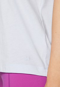 Light gray cotton T-shirt with short sleeves, featuring a small embroidered logo at the hem. Paired with bright purple leggings.
