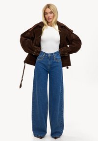 Veste oversized en daim marron avec de grands revers, associée à un jean taille haute à jambes larges bleu clair avec des accents cloutés. Haut ajusté blanc en dessous.