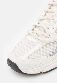 Bílé tenisky s horní částí z prodyšné síťoviny, ozdobami z texturované usně a černou gumovou podrážkou. Mají silné tkaničky a zakřivené švy.