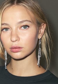 Orecchini pendenti argentati con catene e accenti metallici circolari. Pella liscia con trucco minimale, indossando un top nero.