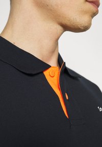 Czarna koszulka polo z pomarańczową listwą guzika. Charakteryzuje się miękką teksturą i klasycznym designem kołnierzyka. Zawiera małe logo na piersi.