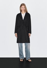 Czarny płaszcz z wełny w stylu tailored, z zapięciem na jeden guzik, klapami o zaokrąglonym kształcie oraz dwoma przednimi kieszeniami, zestawiony z jasnymi dżinsami.