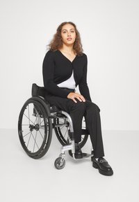 Black cardigan over a white top, seated in a wheelchair. Black pants and shoes with a chain accent. Smooth textures and modern design.