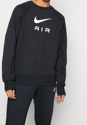 Osoba ubrana w czarną bluzę z kapturem z białym logo Nike i napisem "AIR", w połączeniu z czarnymi dresami Nike z małym logo.