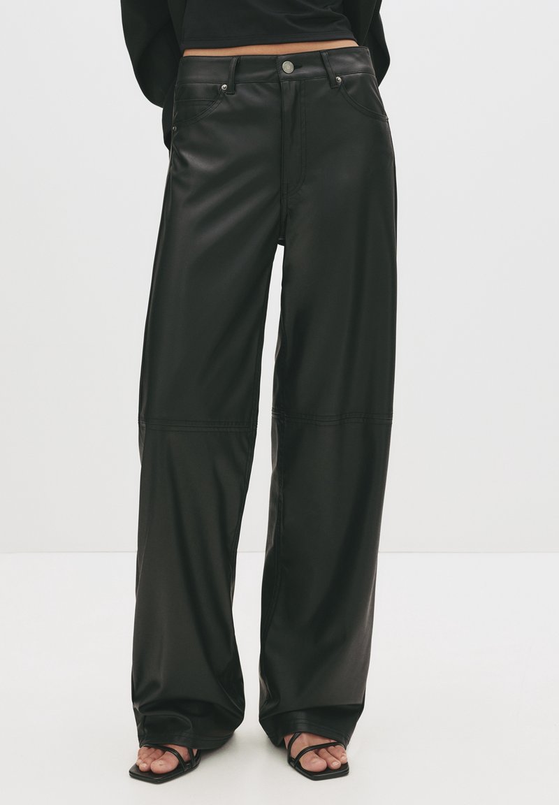 Personne portant un pantalon en cuir noir à taille haute, jambes larges avec poches avant et sandales noires à bout ouvert, debout sur un sol blanc.