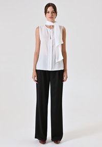 Weiße ärmellose Bluse mit einem Halsband, kombiniert mit schwarzen weit geschnittenen Hosen. Glattes Material, minimalistisches Design und Detail mit Knopfverschluss.