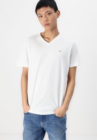 T-shirt en coton blanc avec un col en V et des manches courtes, arborant un petit logo gris sur la poitrine, associé à un jean en denim bleu.