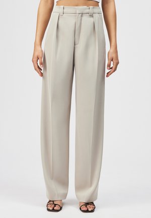 Kobieta w beżowych spodniach z wysokim stanem i szerokimi nogawkami, w czarnych sandałach z paskami i kwadratowym noskiem, stojąca na jednolitym tle.