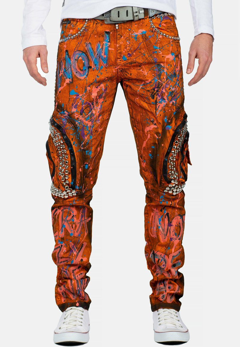 Oranje denim broek met opvallende blauwe en roze spetters, metalen studs en kniezakken met zwarte stoffen accenten.