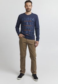 Langarm-Navy-T-Shirt mit auffälligem Text, kombiniert mit beigen Hosen. Schwarze Sneaker mit weißen Sohlen vervollständigen das Outfit.