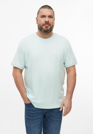 Baardige man met naar achteren gekamd haar draagt een lichtblauw kort mouwenshirt en een blauwe spijkerbroek, staand met één hand in zijn zak tegen een witte achtergrond.