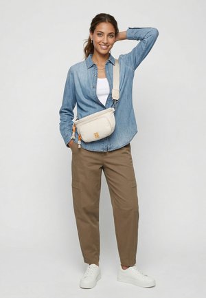 Frau lächelt, trägt ein helles Jeanshemd, weißes Top, braune Cargohosen, weiße Sneaker und eine beige Umhängetasche, steht mit einer Hand am Kopf.