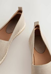 Beige mokkasiner i ruskind med elastiske sideindsatser, syninger og vævede jutedetaljer på en flad sål. Inderforingen er brun.