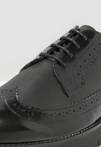 Chaussure noire en cuir avec un travail de brogue complexe, bout rond et laçage traditionnel. Présente des surfaces lisses et texturées.