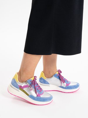 Feet wearing colorful sneakers with blue, silver, pink, yellow, and olive accents, paired with black cropped pants on a white background.