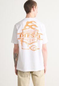 T-shirt bianco in cotone con una grafica arancione e il testo "Diesel" e motivi di fiamme sul retro. Maniche corte e vestibilità ampia.