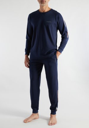 Man in navy blue long-sleeve top and matching joggers with side stripes, standing barefoot against a plain light background.