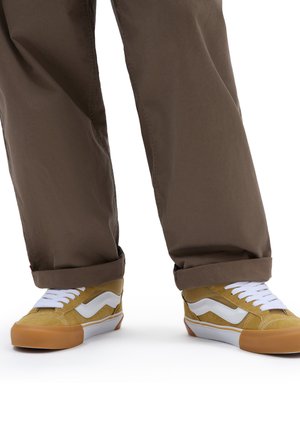 Zapatillas amarillas y blancas con parte superior texturizada, cordones blancos y suela de goma marrón, combinadas con pantalones marrones de corte holgado.