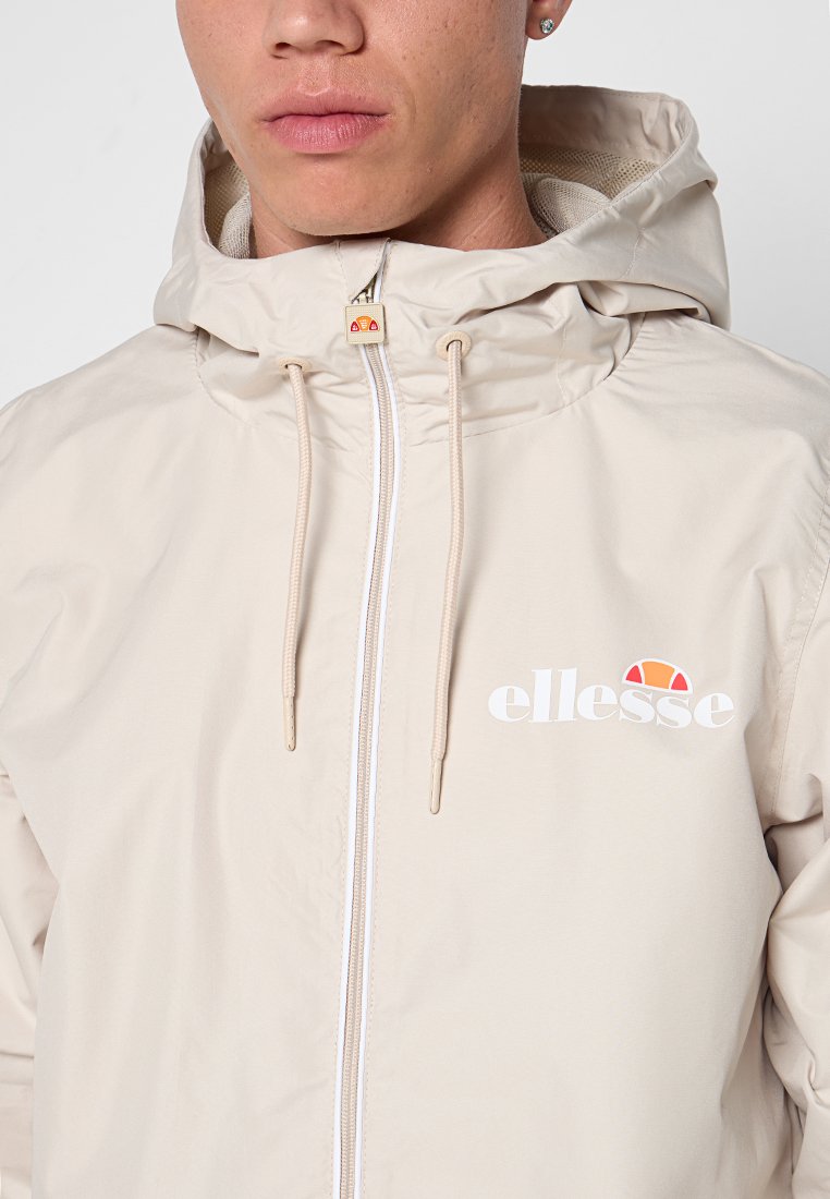 Persona joven vistiendo una chaqueta con capucha beige con cremallera y cordones, que presenta un logo de ellesse en blanco y naranja en el pecho.