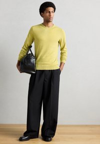 Πράσινο πλεκτό πουλόβερ με στρογγυλή λαιμόκοψη, συνδυασμένο με μαύρο παντελόνι φαρδιάς γραμμής. Το μοντέλο κρατά μια μαύρη υφασμάτινη τσάντα με διπλές λαβές.