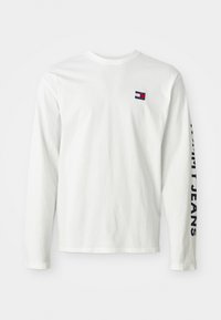 Witte langemouwen overhemd met klein Tommy Jeans logo op de borst en grote Tommy Jeans tekst verticaal gedrukt op de linker mouw.