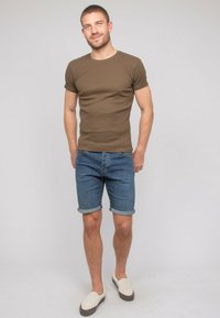 T-shirt marron ajusté à manches courtes, short en denim, manches retroussées, chaussures décontractées à enfiler avec une tige claire et une semelle foncée. Fond neutre.