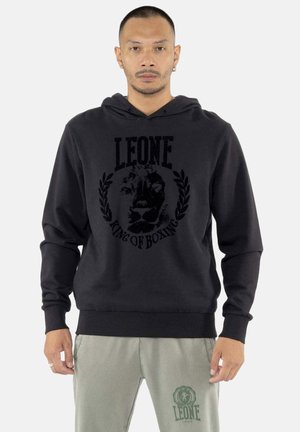 Homme portant un sweat à capuche noir avec le logo « LEONE King of Boxing » et un pantalon gris avec l’emblème « LEONE 1947 » sur la cuisse gauche.