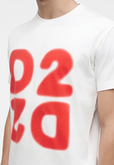 T-shirt blanc à manches courtes avec de grands chiffres et lettres rouges peintes à la bombe 02 3a sur le devant, porté par une personne.